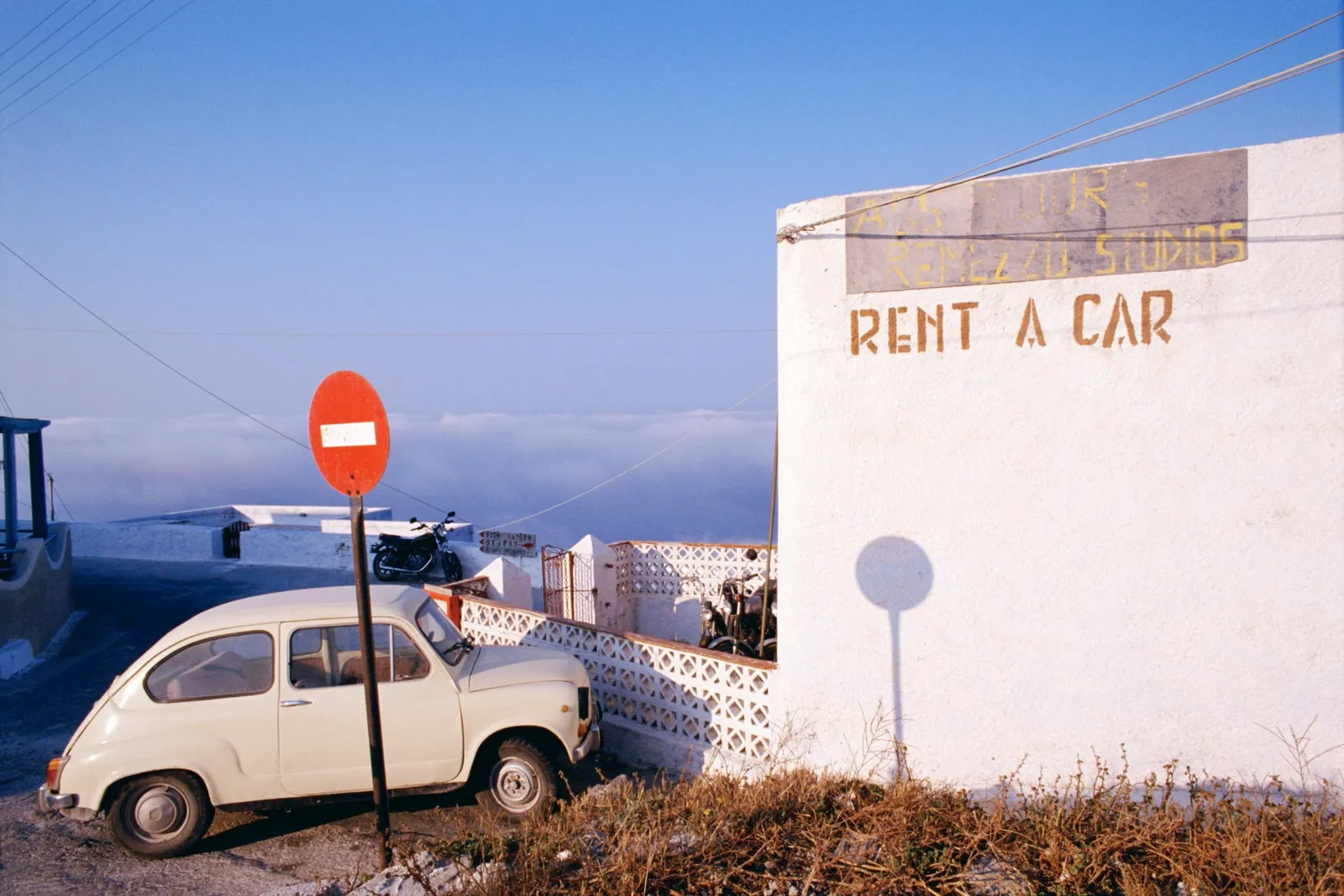 An older style car parked outside of a building with a sign reading ?Rent a Car'; Greece.